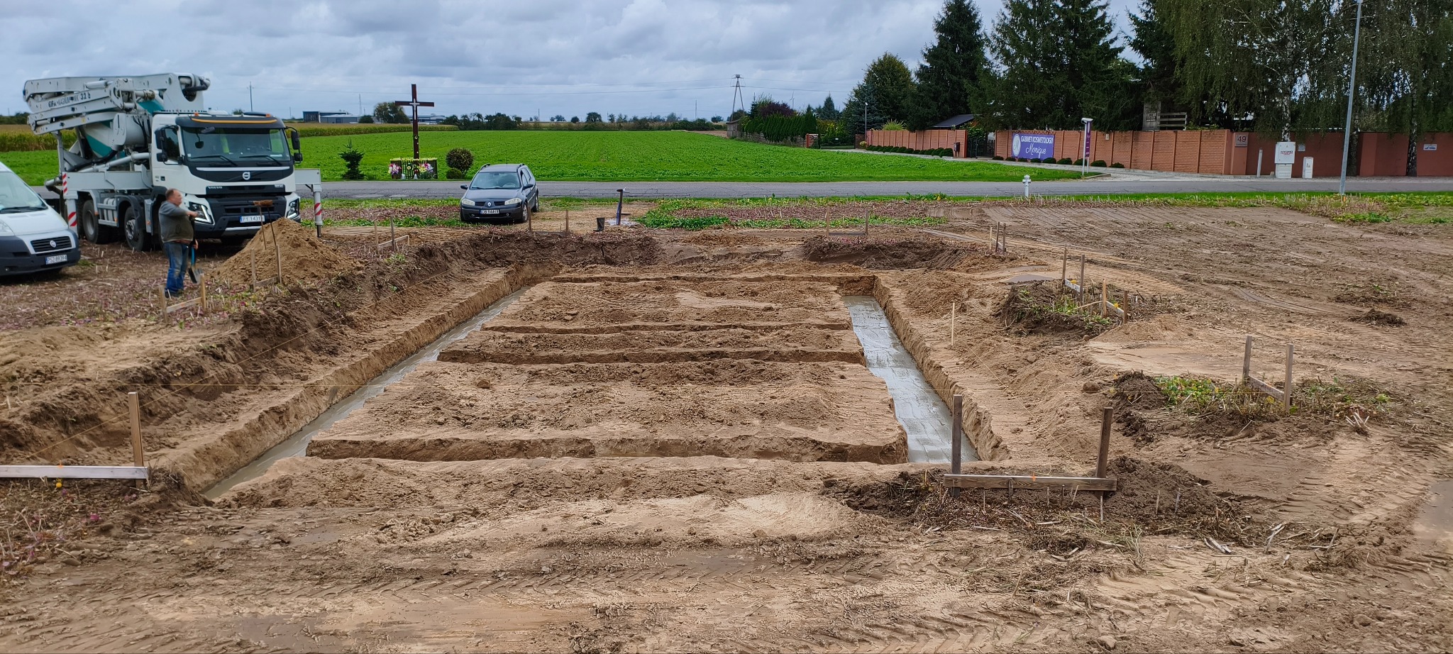 Wykopane i częściowo zalane fundamenty pod budynek, widoczny samochód z pompą do betonu, krzyż przydrożny i krajobraz polny w tle.