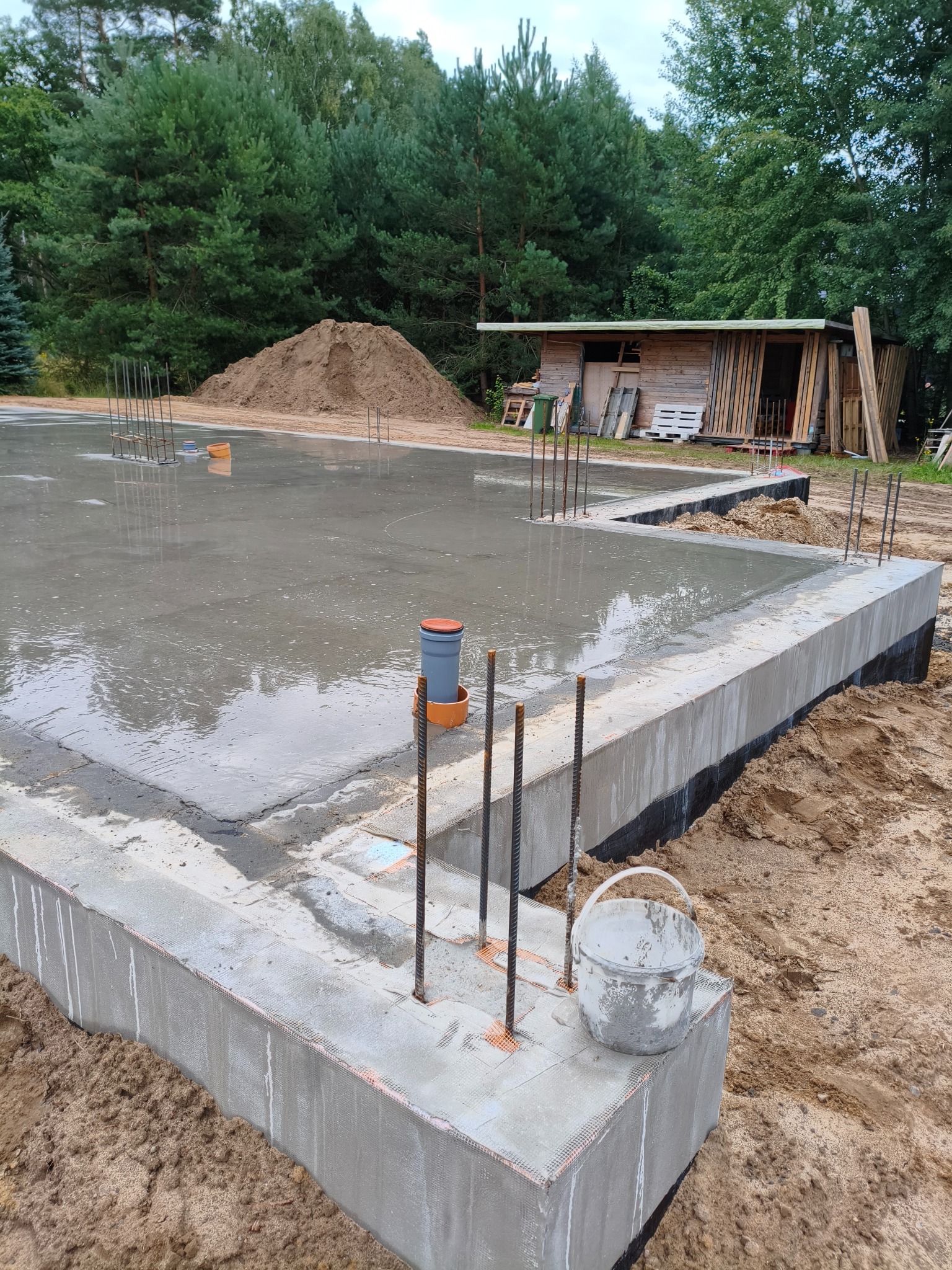 Zalane fundamenty domu z wystającymi prętami zbrojeniowymi, widoczny szalunek, wiadro i sterta piasku w tle, obok drewniana wiata. Ujęcie z bliska, w pochmurny dzień.