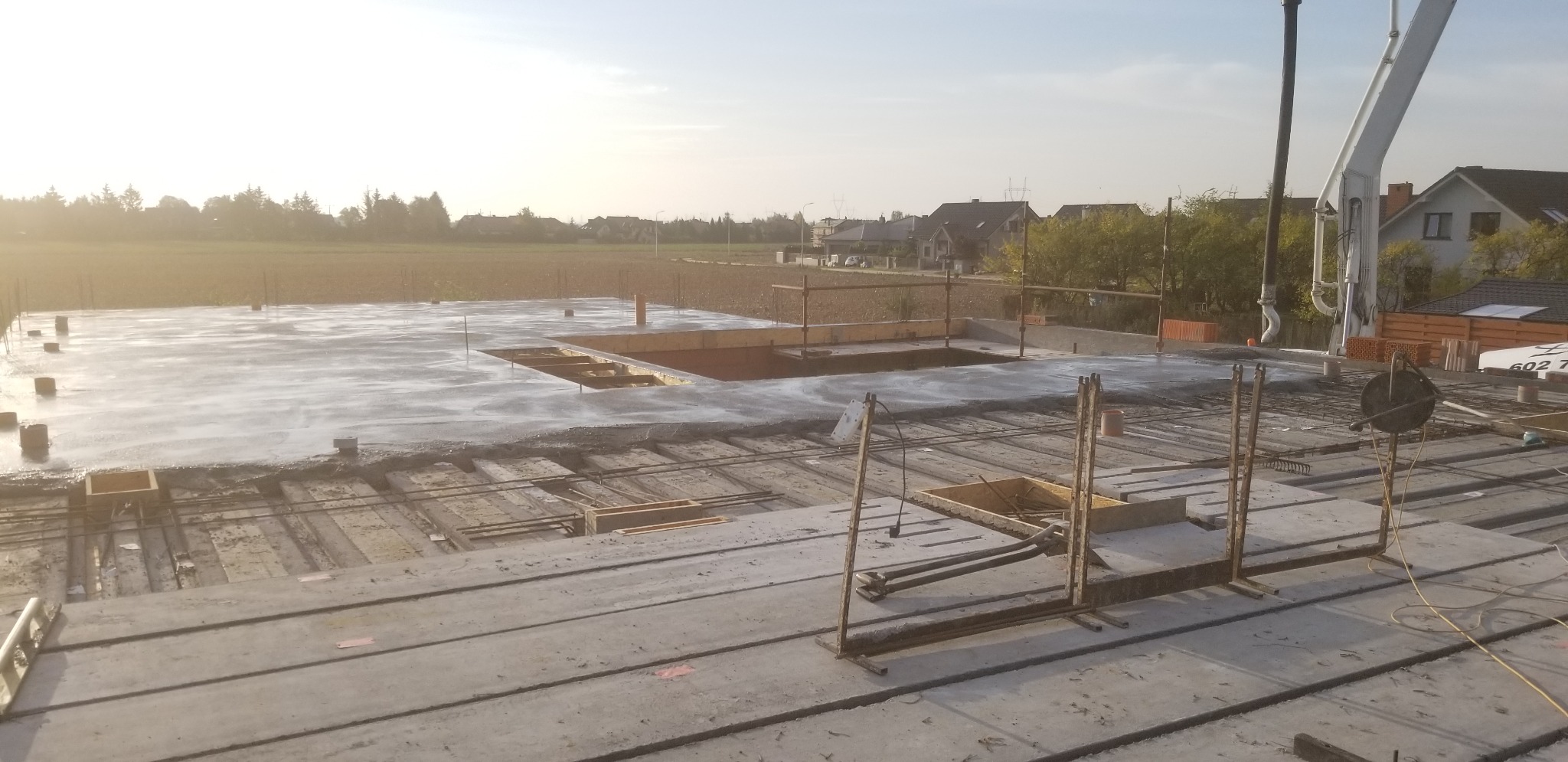 Wylany beton na stropie budynku w trakcie budowy, widoczne zbrojenia i szalunki, w tle krajobraz wiejski i maszyna do podawania betonu.