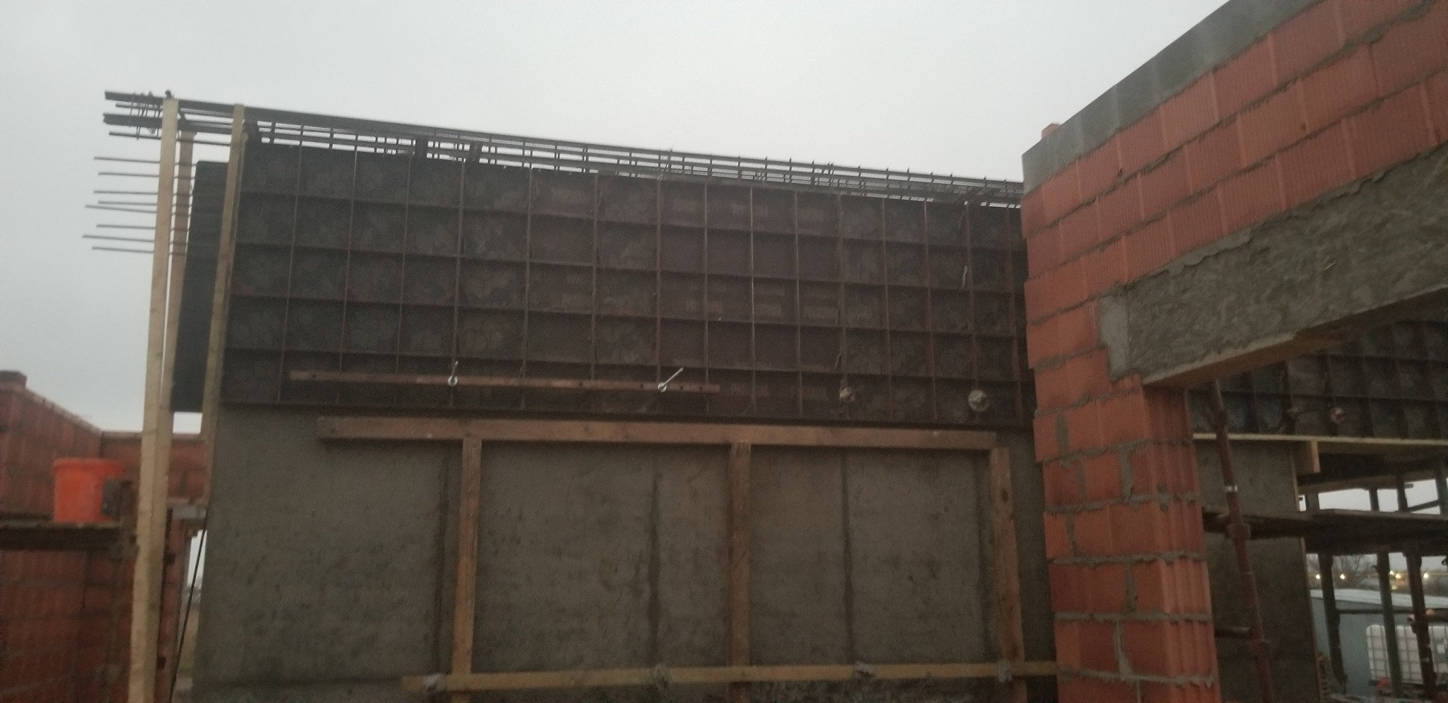 Szare szalunki zbrojeniowe na ścianie budynku w trakcie budowy, widoczny fragment ściany z czerwonej cegły dziurawki, na górze wystające pręty zbrojeniowe, w tle pochmurne niebo.