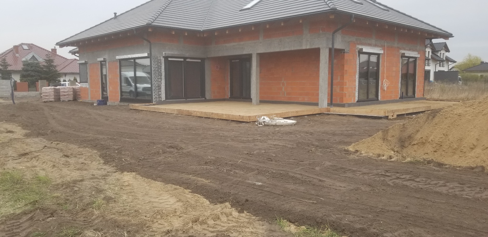Surowy, parterowy dom z czerwonej cegły z szarym dachem i drewnianym tarasem w trakcie budowy, widoczne worki z materiałami budowlanymi i hałda piasku na zewnątrz.