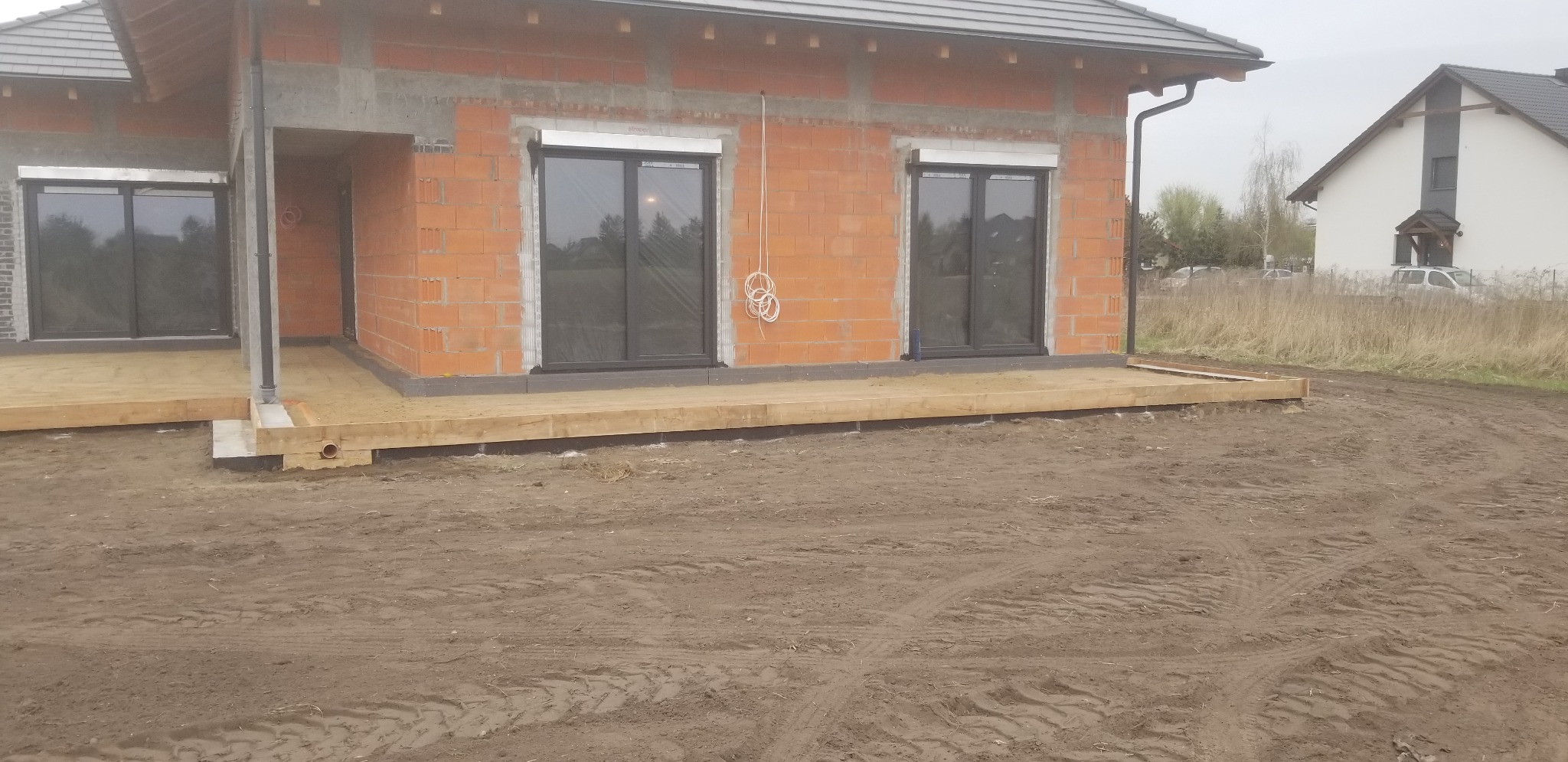 Surowy stan domu jednorodzinnego z czerwonej cegły, widoczne okna z roletami zewnętrznymi, drewniana podkonstrukcja tarasu, teren wokół w stanie przygotowania, drugi dom w tle.