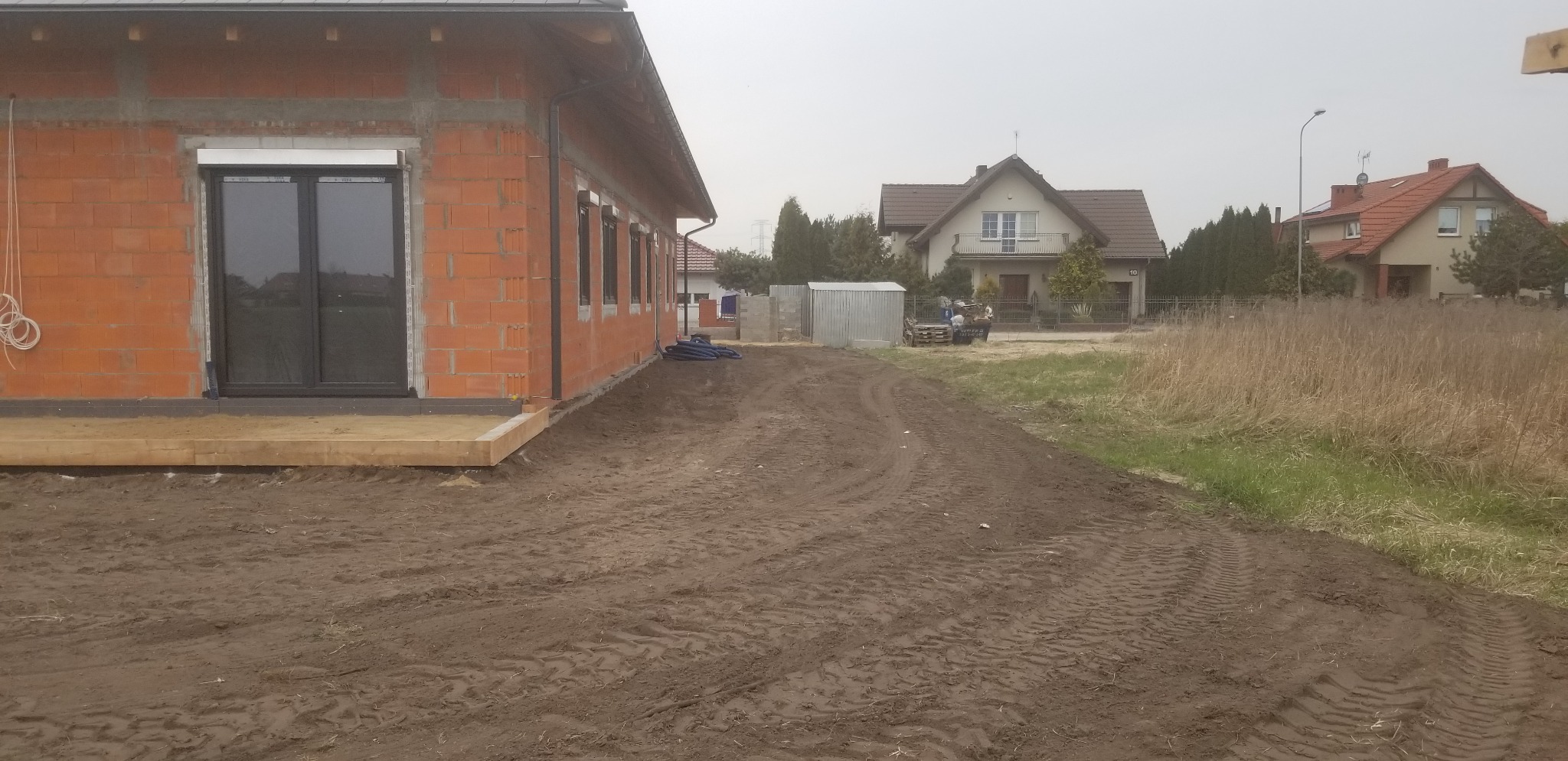 Surowy stan domu jednorodzinnego z czerwonej cegły z zamontowanymi oknami i przygotowanym terenem wokół, widoczne ślady opon na ziemi.