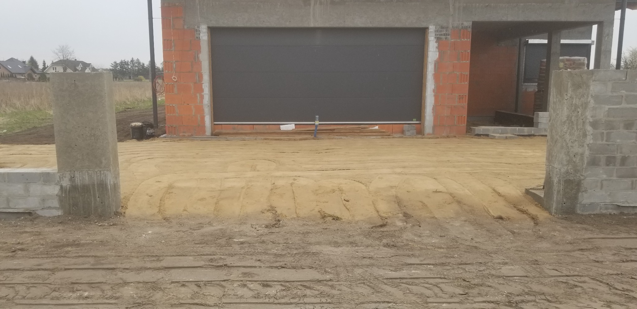 Surowy stan budowy domu jednorodzinnego z garażem: widoczne ściany z czerwonej cegły, szara brama garażowa, betonowe słupy wejściowe i wyrównany teren podjazdowy z widocznymi śladami opon.