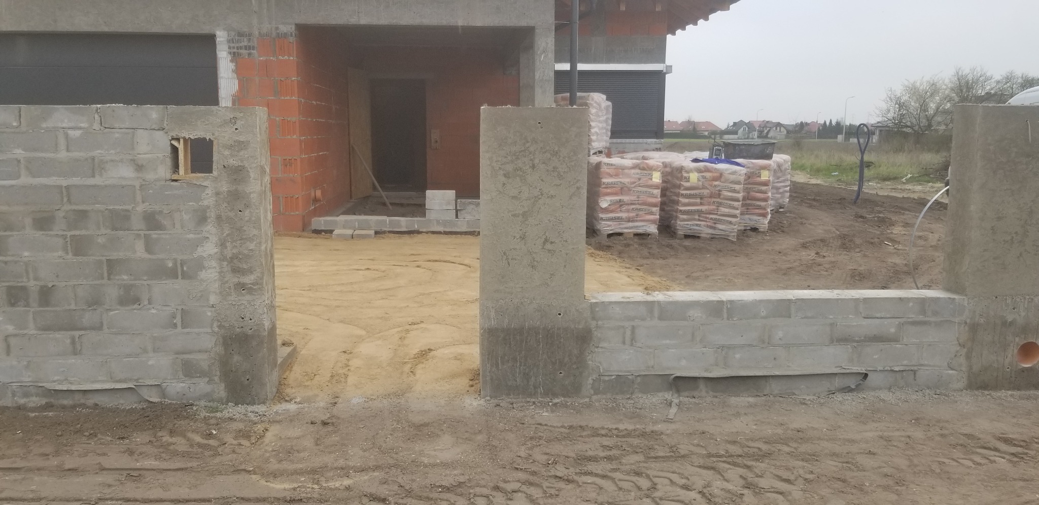 Surowy stan domu w trakcie budowy: widoczne ściany z czerwonej cegły, bloczki betonowe, worki z zaprawą w tle, oraz surowy teren budowy.