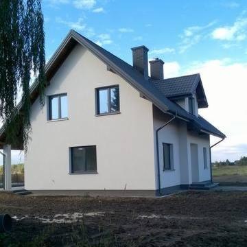 Boczny widok otynkowanego, dwukondygnacyjnego domu murowanego z ciemną dachówką i szarymi ramami okiennymi, z widocznym fragmentem rynny i komina, na tle błękitnego nieba i pola.