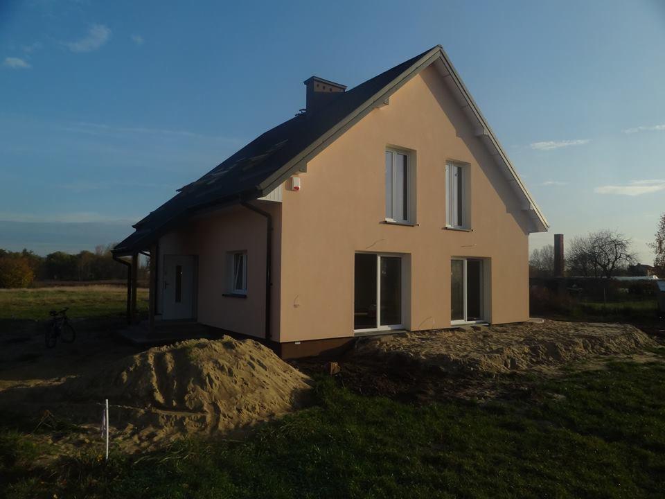 Dwukondygnacyjny dom z beżową elewacją i ciemnym dachem, widoczny wczesną jesienią, z stertą piasku przed budynkiem.