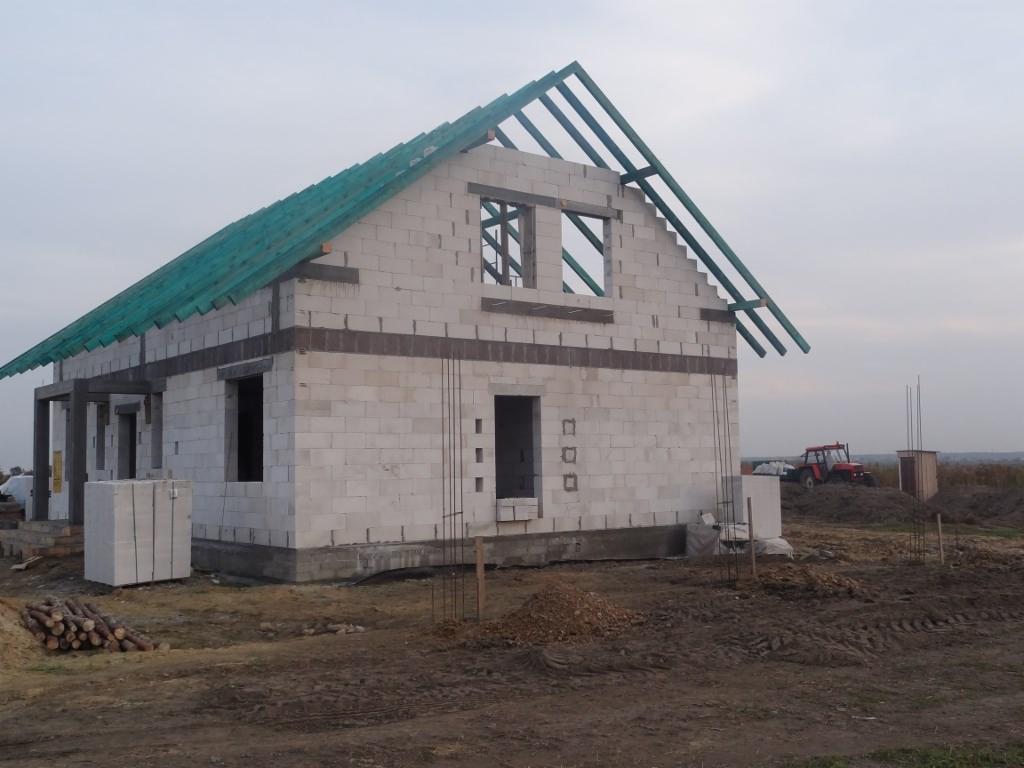 Niedokończony dom z białego pustaka, widoczne drewniane elementy konstrukcji dachu i sterta pustaków obok, w tle traktor.