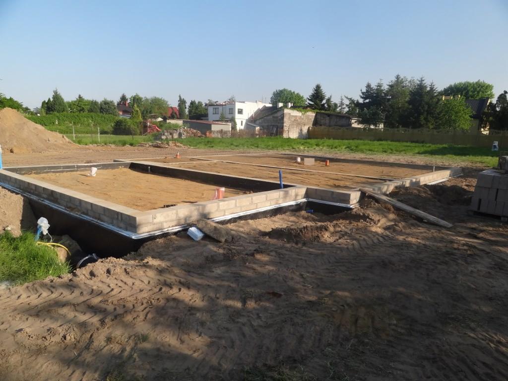 Wylane fundamenty pod dom jednorodzinny z widocznymi bloczkami betonowymi, izolacją poziomą i zasypanym piaskiem wewnątrz, widok z poziomu gruntu na placu budowy.