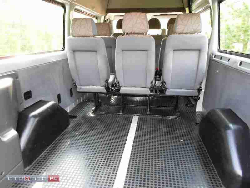 Wnętrze busa z trzema rzędami foteli w odcieniach szarości, widok z tyłu pojazdu na siedzenia z wzorzystymi zagłówkami i podłogę z antypoślizgową fakturą.