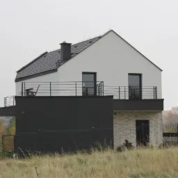 Dwupiętrowy dom jednorodzinny z płaskim dachem, czarną elewacją części parterowej i balkonem z metalową balustradą, widziany z perspektywy trawnika.