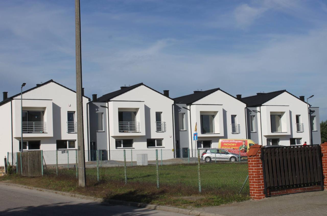 Szereg identycznych, nowoczesnych, dwukondygnacyjnych domów jednorodzinnych z balkonami, w stanie deweloperskim, z czarnymi dachami i białymi elewacjami, ogrodzone siatką. Na drugim planie...