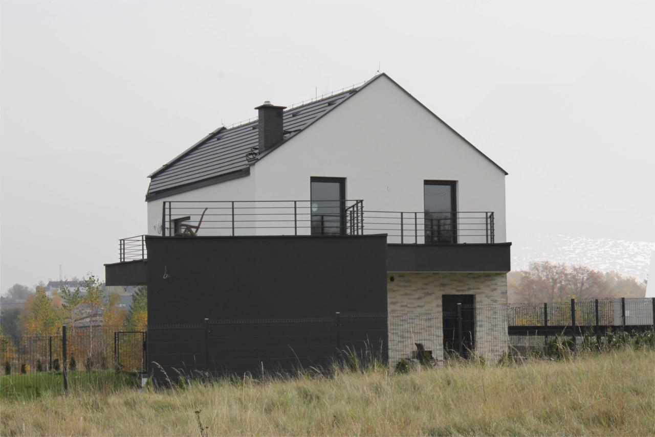 Dwupiętrowy dom jednorodzinny z płaskim dachem, czarną elewacją części parterowej i balkonem z metalową balustradą, widziany z perspektywy trawnika.