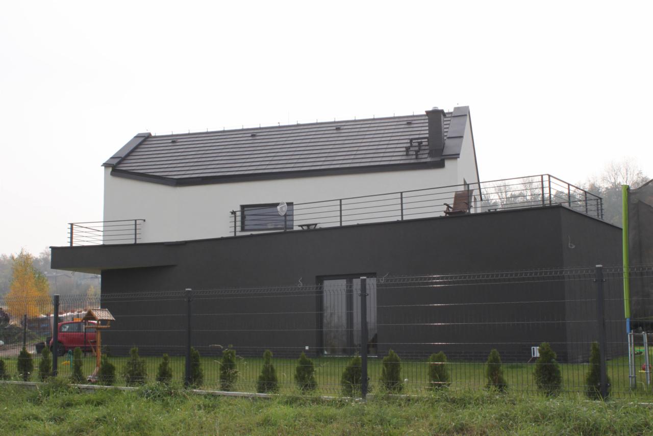 Nowoczesny dom z płaskim dachem, balkonem z metalową balustradą i elewacją w kolorze białym i grafitowym, otoczony niskim żywopłotem z tuj.