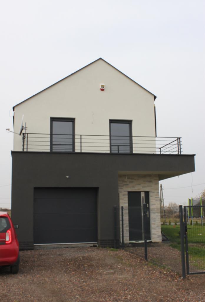 Dwukondygnacyjny dom z garażem i balkonem z czarną balustradą, widok z zewnątrz w pochmurny dzień, z fragmentem czerwonego samochodu po lewej stronie.