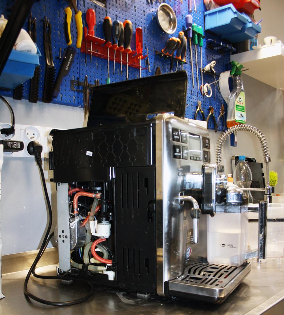 Otwarty ekspres do kawy z widocznymi wewnętrznymi przewodami i elementami, na tle ściany warsztatowej z narzędziami.