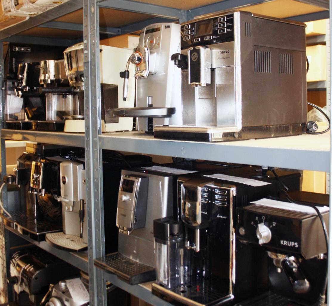Różne modele ekspresów do kawy, marek Saeco i Krups, ustawione na metalowych półkach magazynowych.