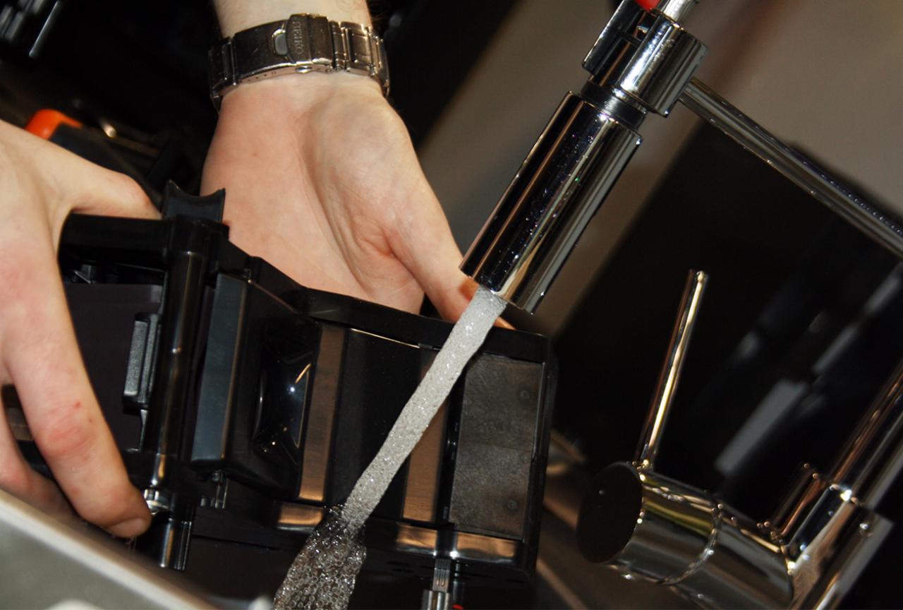 Czyszczenie elementu ekspresu do kawy pod bieżącą wodą, widoczny srebrny kran i fragment dłoni z metalowym zegarkiem.