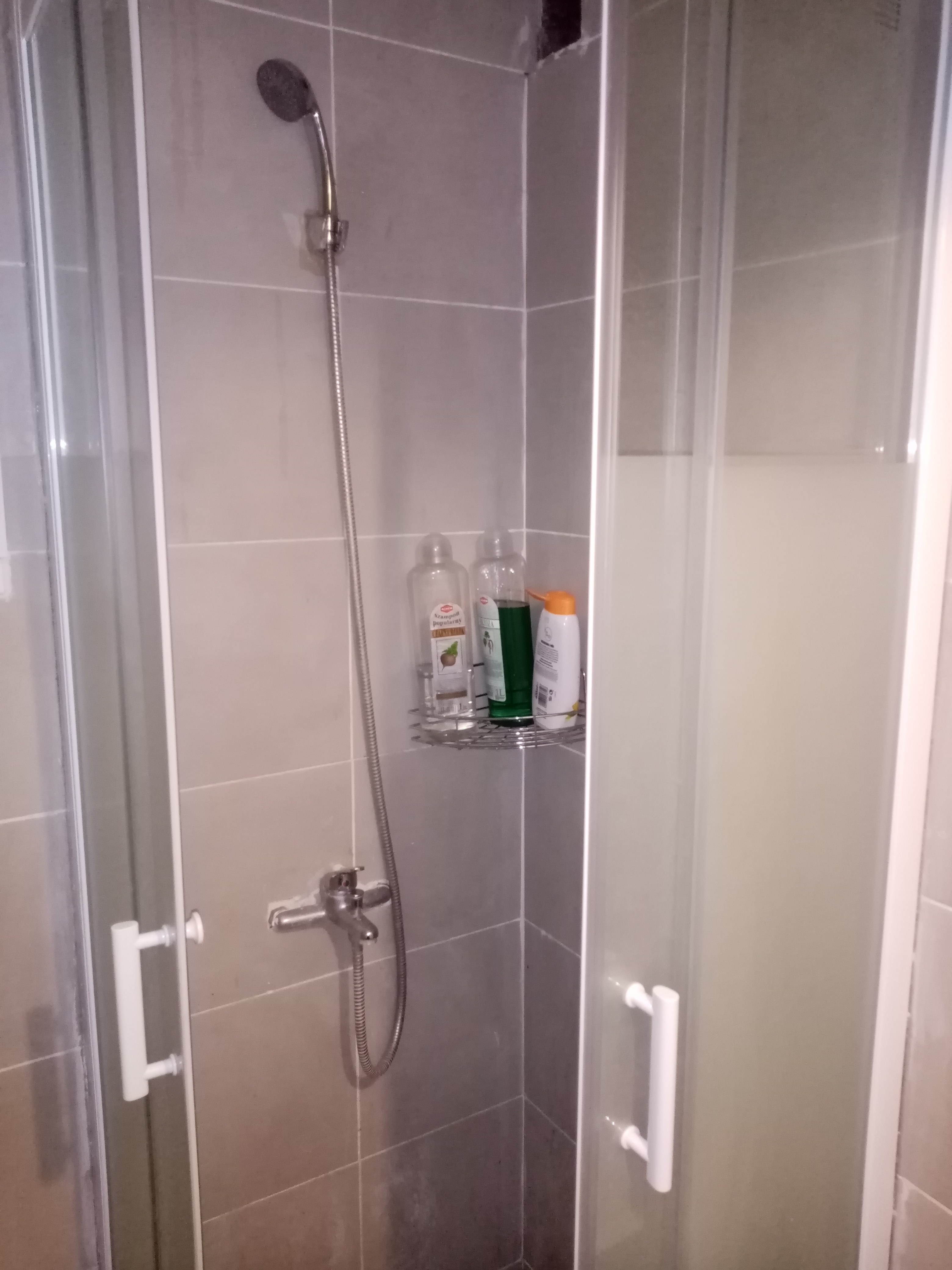 Szklana kabina prysznicowa z chromowaną armaturą i półką na kosmetyki, widoczne szare płytki ścienne z fugami.