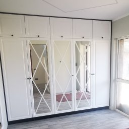 Greko Grzegorz Komorowski - Biała szafa wnękowa z lustrami w romby, czarne obramowanie i gałki, widoczne niedokończone ściany w tle.