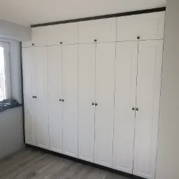 Biała szafa wnękowa z czarnymi akcentami, obejmująca całą ścianę od podłogi do sufitu, z rzędem szafek górnych, widoczna na tle jasnych paneli podłogowych.