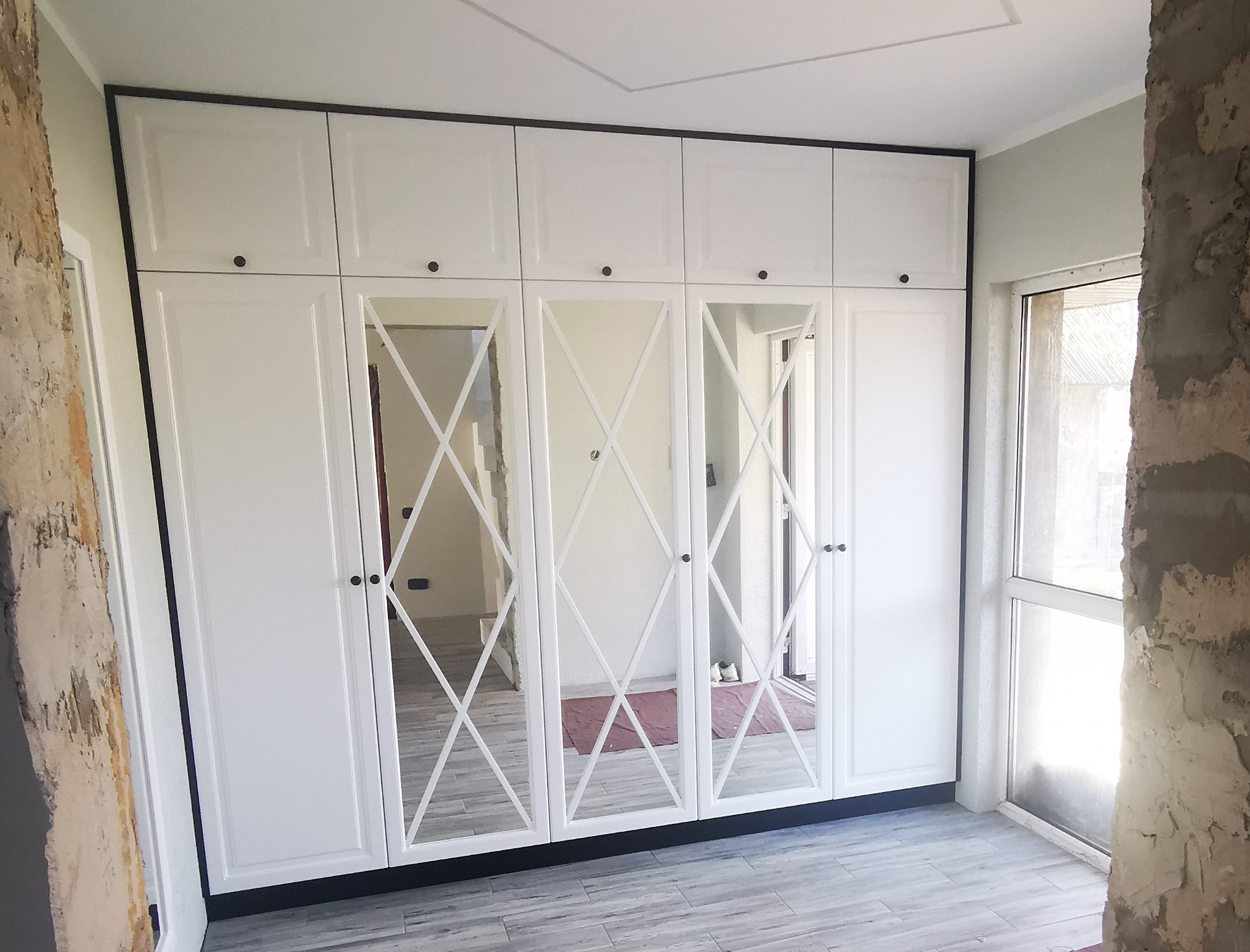 Biała szafa wnękowa z lustrami w romby, czarne obramowanie i gałki, widoczne niedokończone ściany w tle.