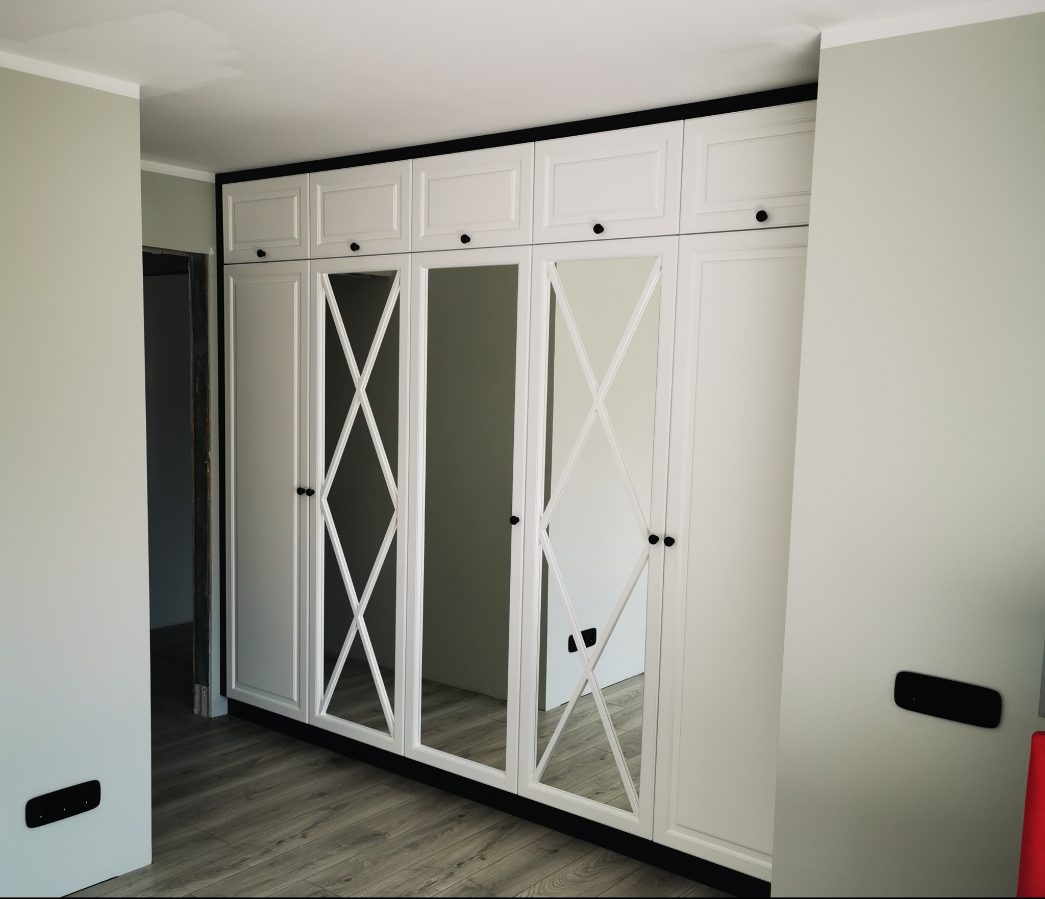 Biała szafa wnękowa z lustrami i czarnymi detalami, widoczna na tle szarej ściany i drewnianej podłogi, z fragmentem ościeżnicy drzwiowej po lewej stronie.