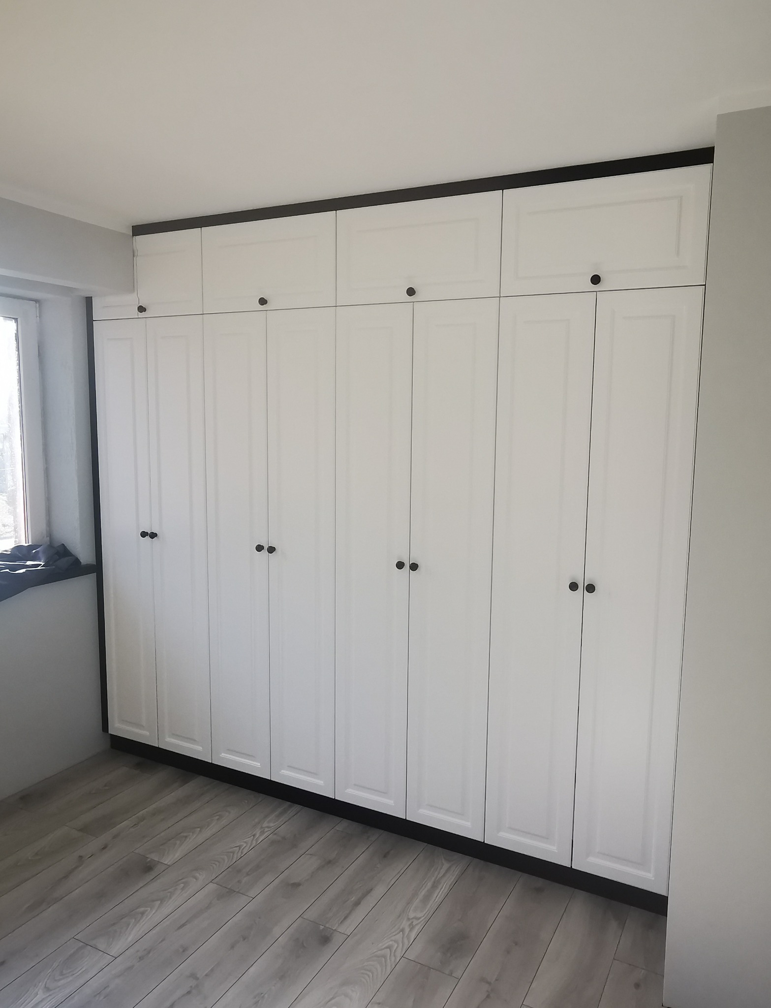 Biała szafa wnękowa z czarnymi akcentami, obejmująca całą ścianę od podłogi do sufitu, z rzędem szafek górnych, widoczna na tle jasnych paneli podłogowych.