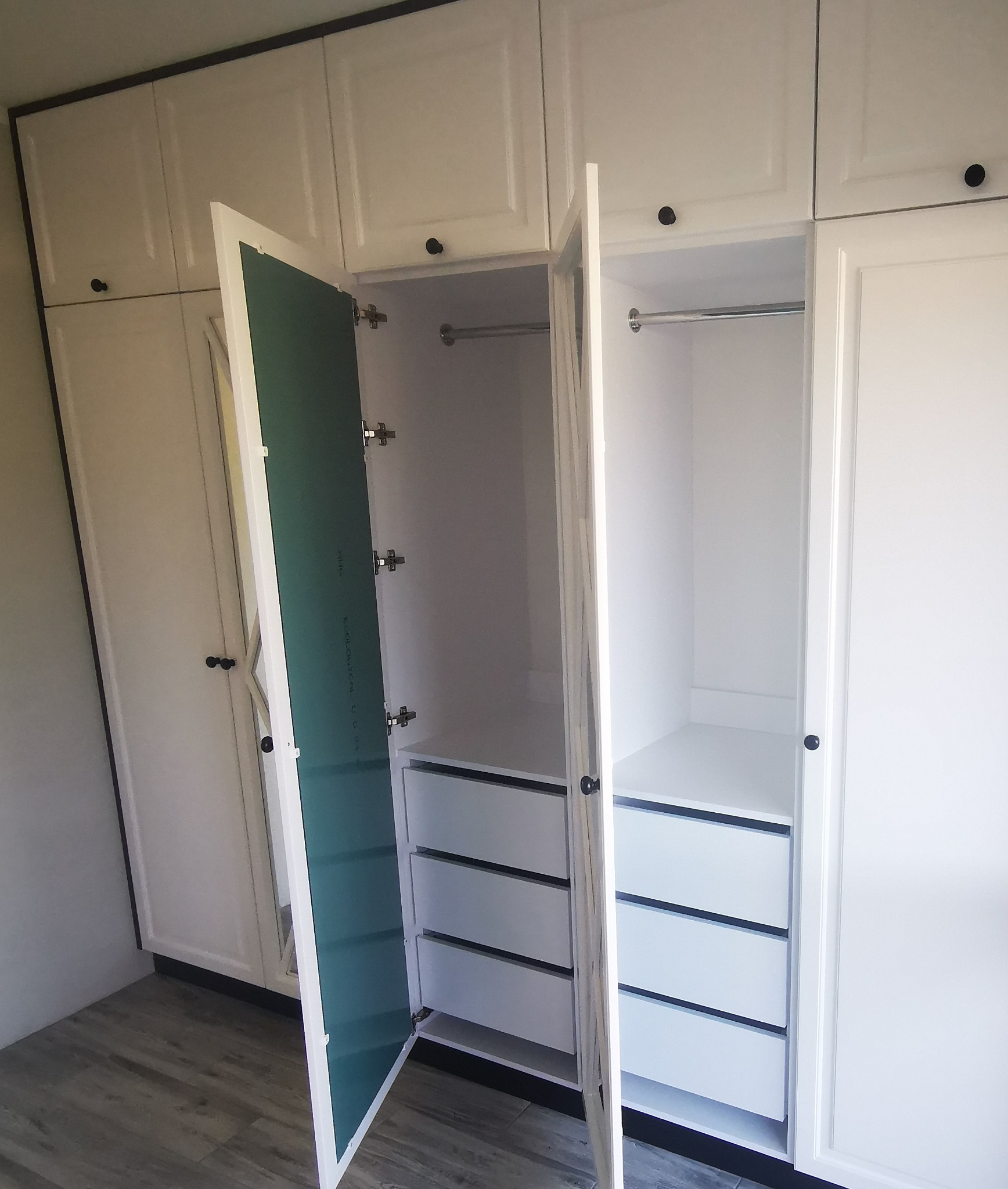 Biała szafa wnękowa z otwartymi drzwiami, ukazująca trzy szuflady i drążek na ubrania, z widoczną zieloną płytą na wewnętrznej stronie drzwi.