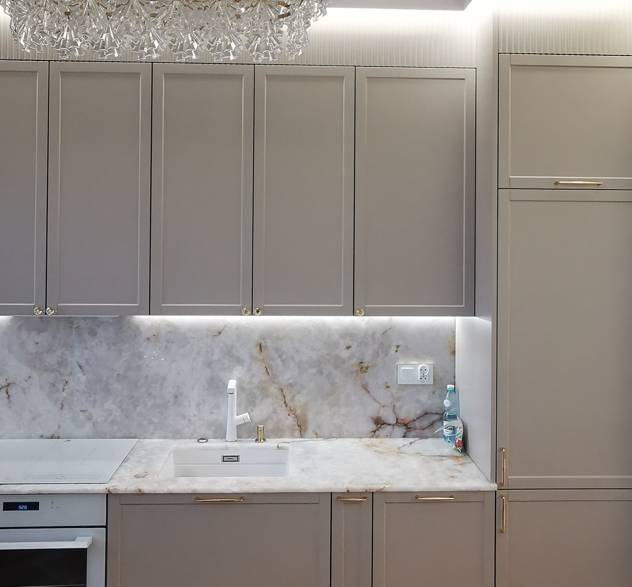 Elegancka kuchnia w odcieniach szarości z marmurowym blatem i kryształowym żyrandolem. Uwagę przykuwają złote uchwyty i subtelne oświetlenie LED pod szafkami.
