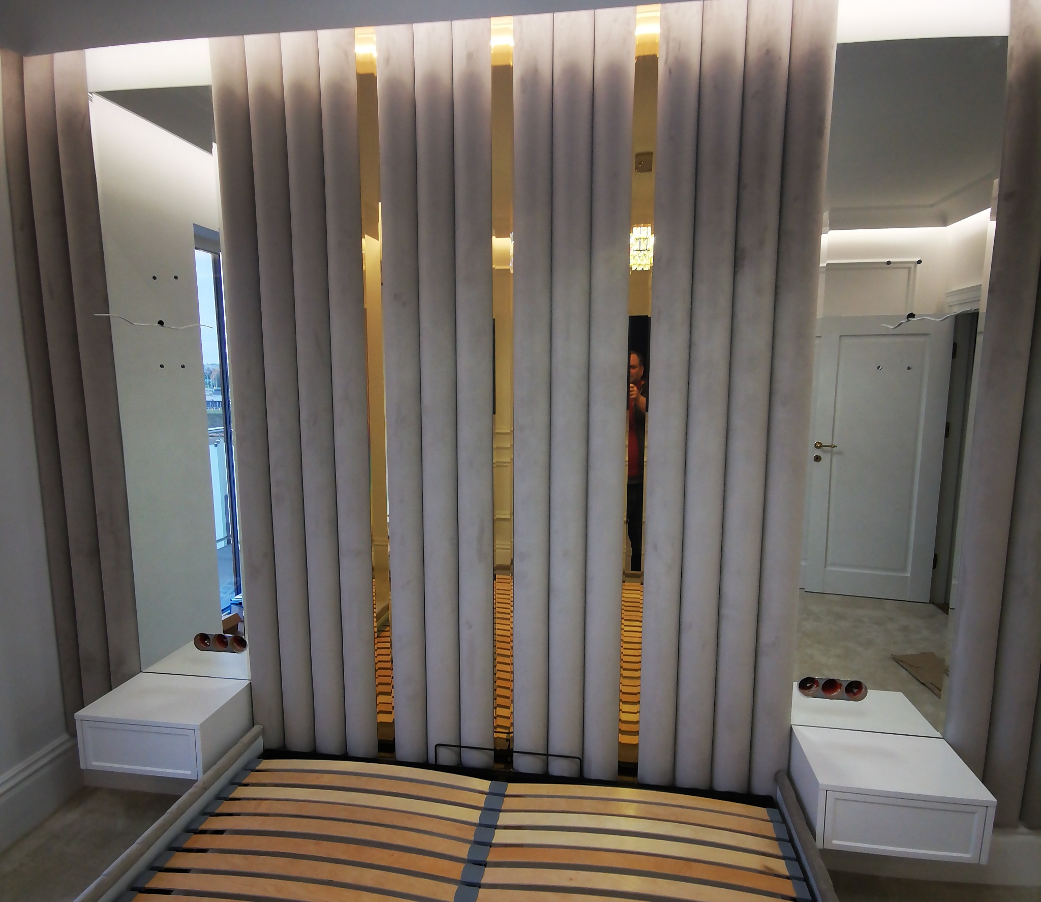 Nowoczesne wezgłowie łóżka z tapicerowanymi panelami w odcieniach szarości, zintegrowane z szafkami nocnymi i lustrami, tworzące spójną aranżację sypialni.