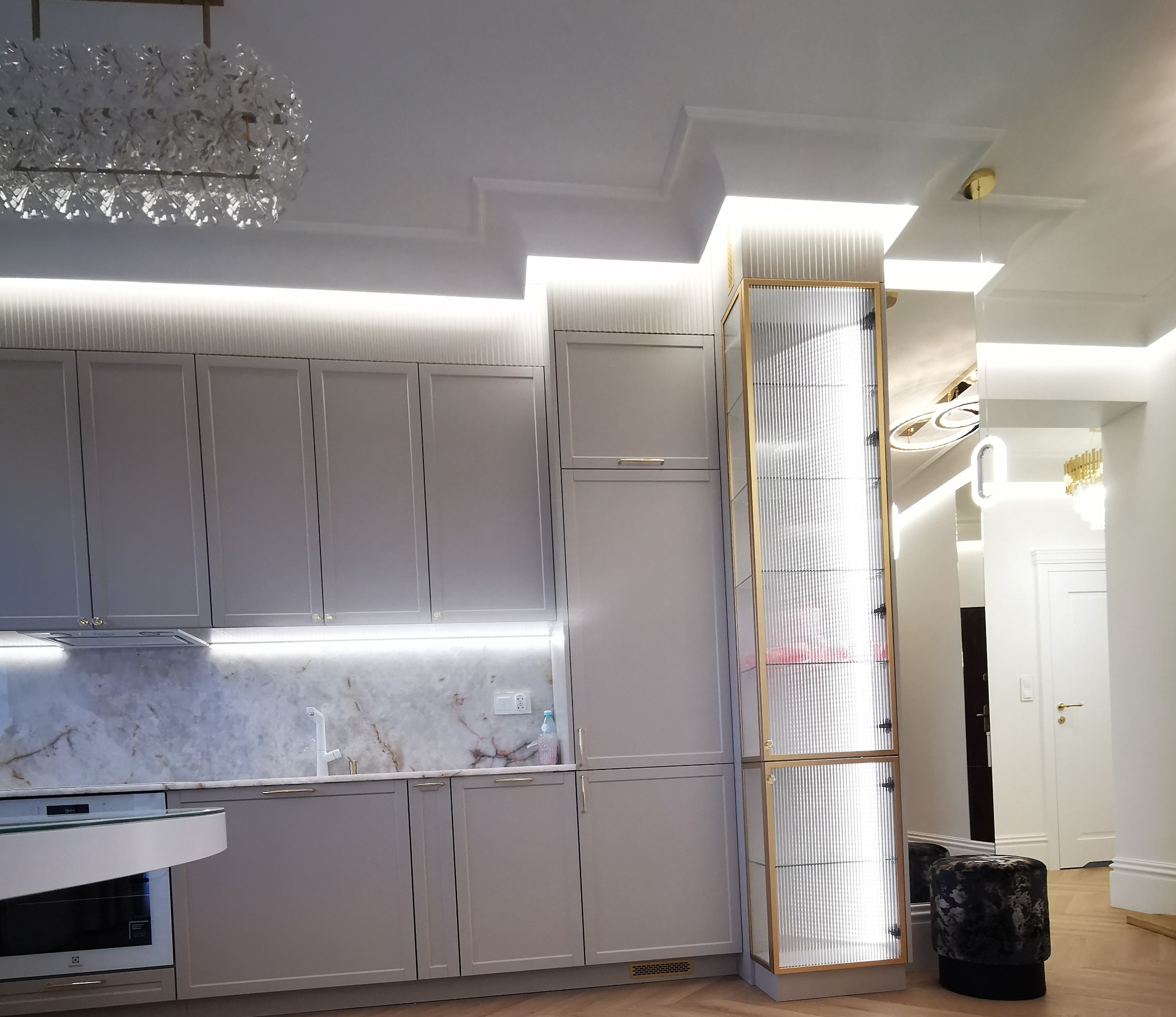 Elegancka kuchnia w odcieniach szarości z marmurowym blatem i oświetleniem LED. Na pierwszym planie przeszklona witryna z złotymi ramami i dekoracyjny puf.