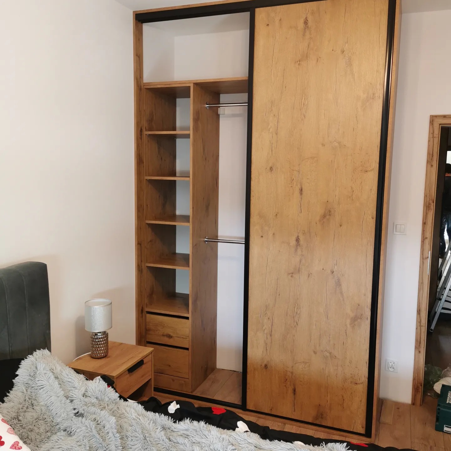 Wnętrze sypialni z dużą szafą przesuwną o drewnianym wykończeniu i czarnych obramowaniach, obok szafki nocnej z lampką i łóżka z puszystą narzutą.