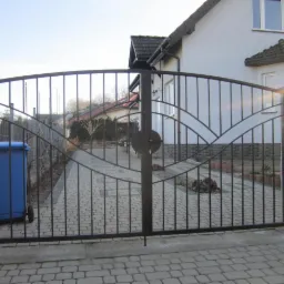 Metalowa brama wjazdowa z dekoracyjnymi łukami, osadzona na kamiennych słupkach. Widoczny podjazd z kostki brukowej i fragment domu z oknami.
