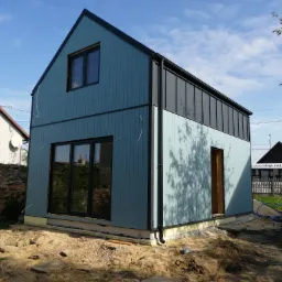 Wznoszony, dwukondygnacyjny budynek w stylu nowoczesnej stodoły, z niebieską elewacją z desek i czarnymi elementami wykończeniowymi, widoczny na etapie wylewania fundamentów i prac ziemnych.