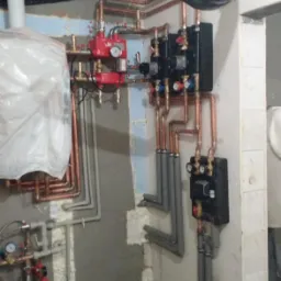 Instalacja centralnego ogrzewania z miedzianymi rurami, czarnymi skrzynkami sterującymi i czerwonymi pompami obiegowymi na tle surowej ściany przed wykończeniem.