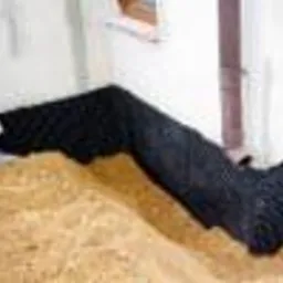 Czarna perforowana rura ułożona w wykopie przy fundamencie budynku, zabezpieczona geowłókniną. Widoczny fragment ściany piwnicy i rura spustowa.