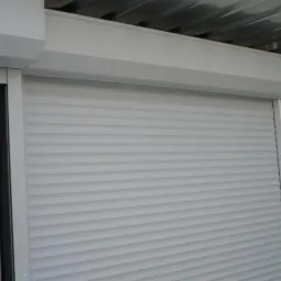 Zamknięta, biała osłona okienna z poziomymi lamelami, widoczna rama okna oraz metalowa konstrukcja stropu.