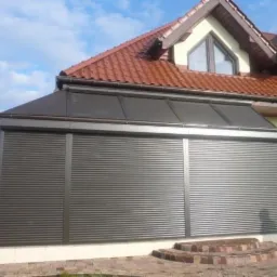 Zewnętrzne, aluminiowe osłony okienne w kolorze grafitowym, zamontowane na przeszklonej werandzie przylegającej do domu z czerwoną dachówką.