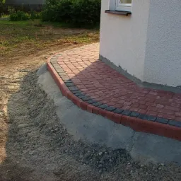 Ceglana ścieżka z czarnym obramowaniem, przylegająca do białej ściany budynku, otoczona ziemią i trawą. Widok z boku, ciepłe światło.