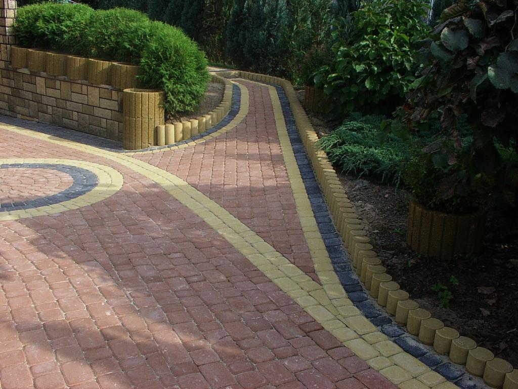 Kaskadowa ścieżka z kostki brukowej w odcieniach czerwieni, żółci i szarości, obrzeża z palisad. Elementy małej architektury ogrodowej i roślinność w tle.