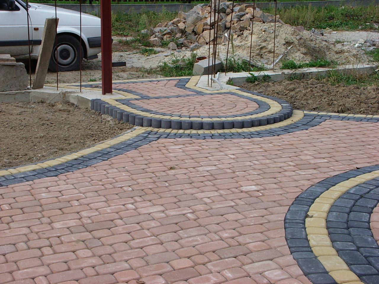 Ułożona kostka brukowa w odcieniach czerwieni, szarości i żółci tworzy ścieżkę z dekoracyjnymi obrzeżami. W tle widoczny samochód i sterta kamieni.