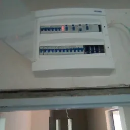 Rozdzielnica elektryczna natynkowa z widocznymi bezpiecznikami i jednym czerwonym wskaźnikiem, zamontowana na ścianie nad futryną drzwi w surowym stanie.