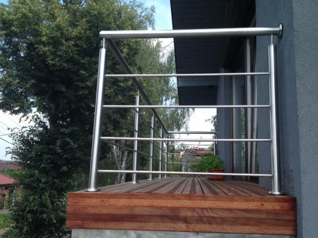 Balkon z balustradą ze stali nierdzewnej, drewniana podłoga z desek kompozytowych, widok na zieleń i fragment budynku.