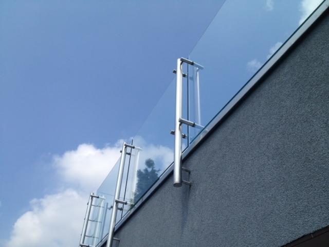 Szklana balustrada balkonowa z metalowymi słupkami, zamontowana na elewacji budynku, widok z dołu na tle błękitnego nieba z chmurami.