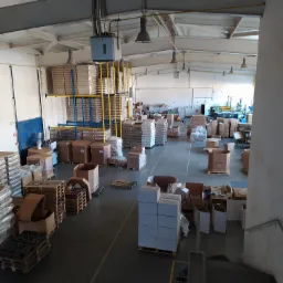 Wnętrze hali produkcyjnej z paletami, kartonami i regałami wypełnionymi produktami, widok z g&oacute;ry z perspektywy schod&oacute;w, surowy industrialny klimat.