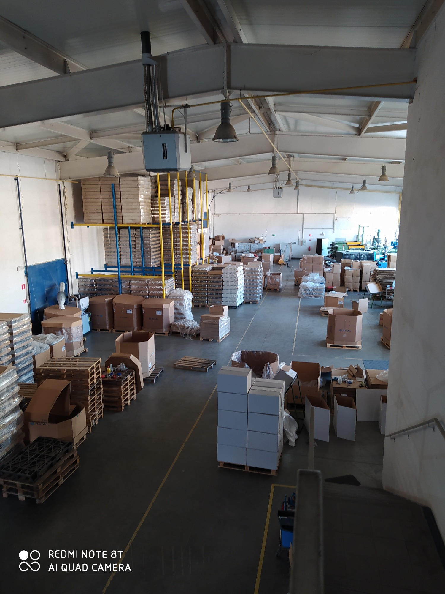 Wnętrze hali produkcyjnej z paletami, kartonami i regałami wypełnionymi produktami, widok z góry z perspektywy schodów, surowy industrialny klimat.