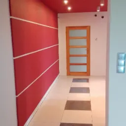 Pomieszczenie po remoncie: czerwona ściana z białymi pasami, drewniane drzwi z szybami, płytki podłogowe w szachownicę, sufit pomalowany na czerwono z zamontowanymi punktowymi lampami.