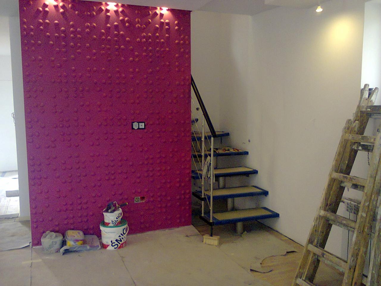 Wnętrze w trakcie remontu: ściana z dekoracyjną fakturą w odcieniach fuksji, częściowo pomalowana ściana, metalowe schody z niebieskimi akcentami i drewniana drabina oparta o ścianę, widoczne...