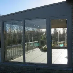 Zewnętrzny widok przeszklonego ogrodu zimowego z przesuwnymi drzwiami, szarym profilem i widocznym fragmentem muru z cegły po prawej stronie.